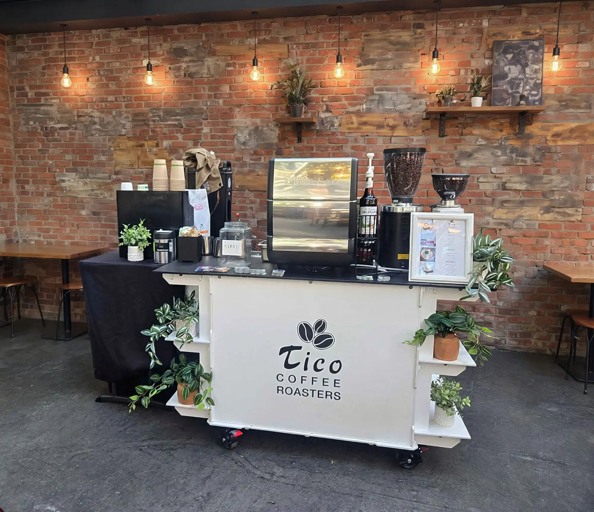 White mobile coffee cart with black wheels and shelves, featuring the ’tico coffee roasters’ logo in black lettering above three stylized coffee bean icons.