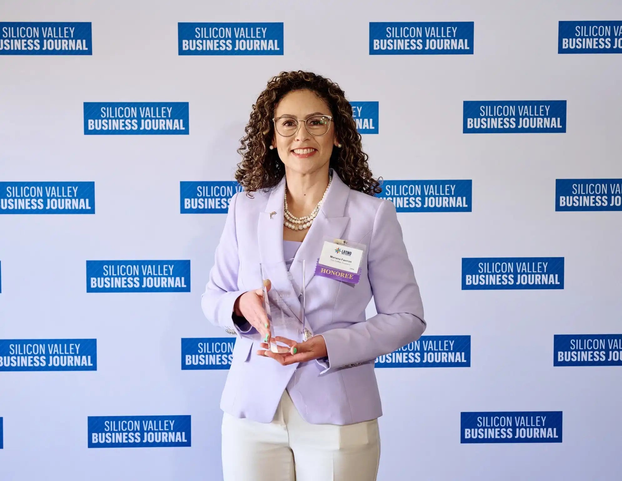 A woman with curly brown hair, wearing glasses, a light purple blazer over a matching top, and white pants, holding a clear award.